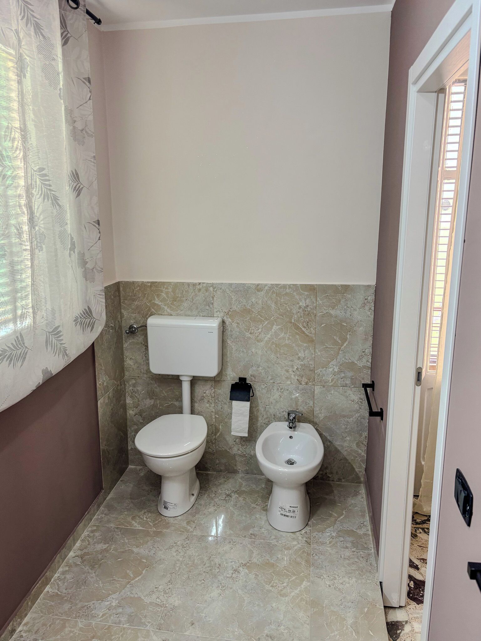 Et badeværelse med toilet, bidet, marmorfliser og et gardin med bladmotiv.