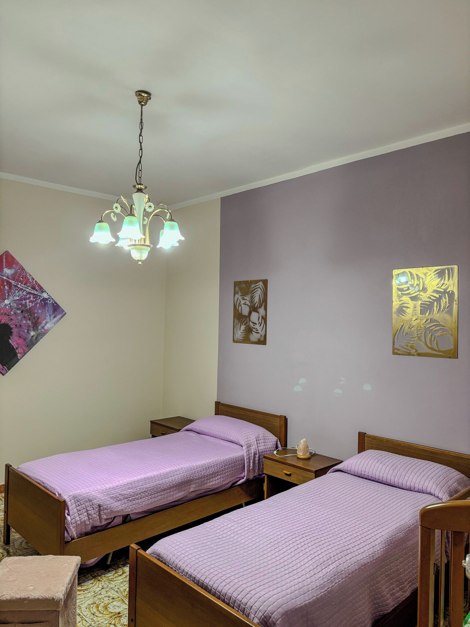 Une chambre avec deux lits simples, une tête de lit en bois, une table de chevet, une petite table, une chaise, un luminaire suspendu et deux œuvres d’art murales.