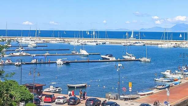 Ein Yachthafen mit mehreren Schiffen, eine Straße direkt am Wasser und im Hintergrund freie Sicht auf das Meer und die Berge.