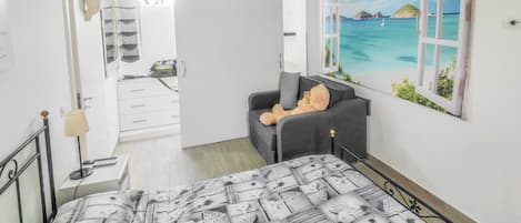 A bedroom with a bed, a grey sofa, a painting of a tropical scene, and a door leading to a bathroom.