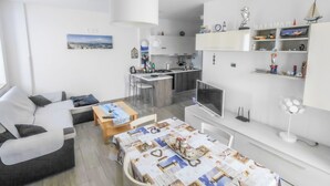 Ein modernes Wohnzimmer mit Sofa, Esstisch mit dekorativer Tischdecke, Küchenbereich mit Spüle und Fernseher sowie ein Bücherregal mit verschiedenen Gegenständen.