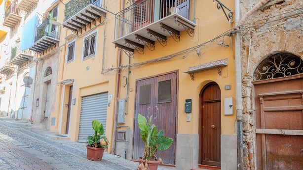 A narrow cobblestone street lined with historic buildings, featuring balconies, wooden doors, and potted plants. - La Discesa Di San Giuseppe (Termini Imerese)
