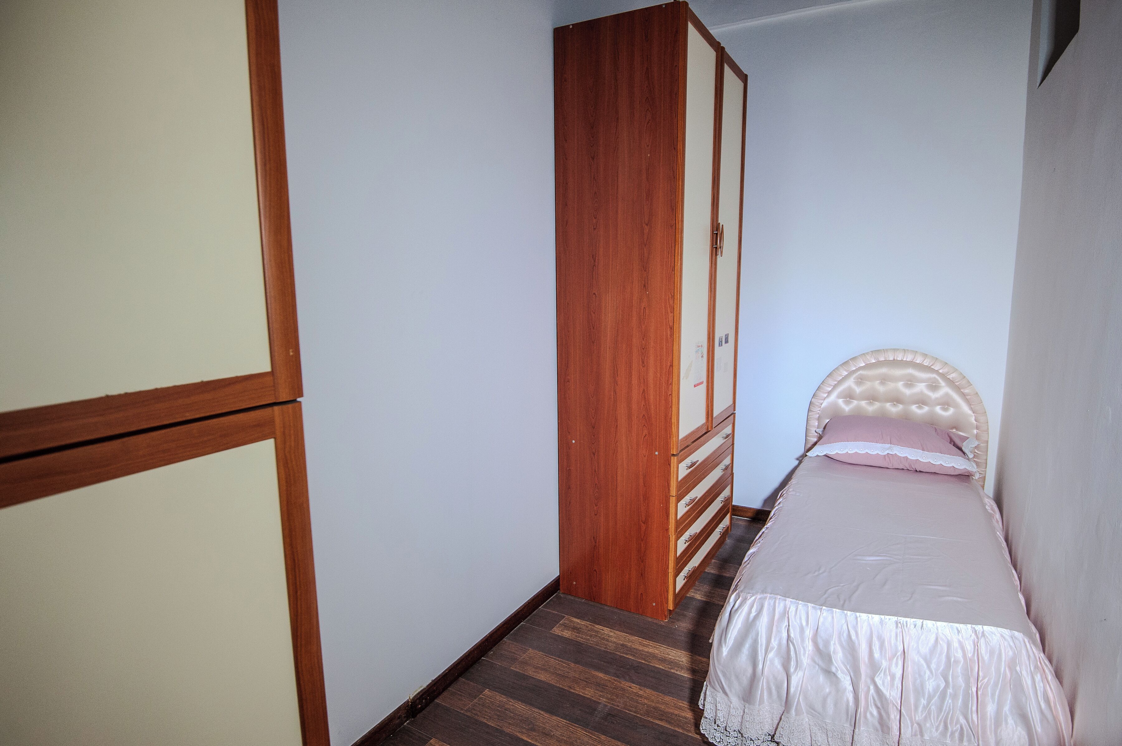 A narrow room with a wooden wardrobe, a bed with a white headboard, and a wooden floor.