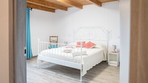 A bedroom with a white metal bed frame, a wooden ceiling, a small bedside table with a lamp, and a chair against the wall.