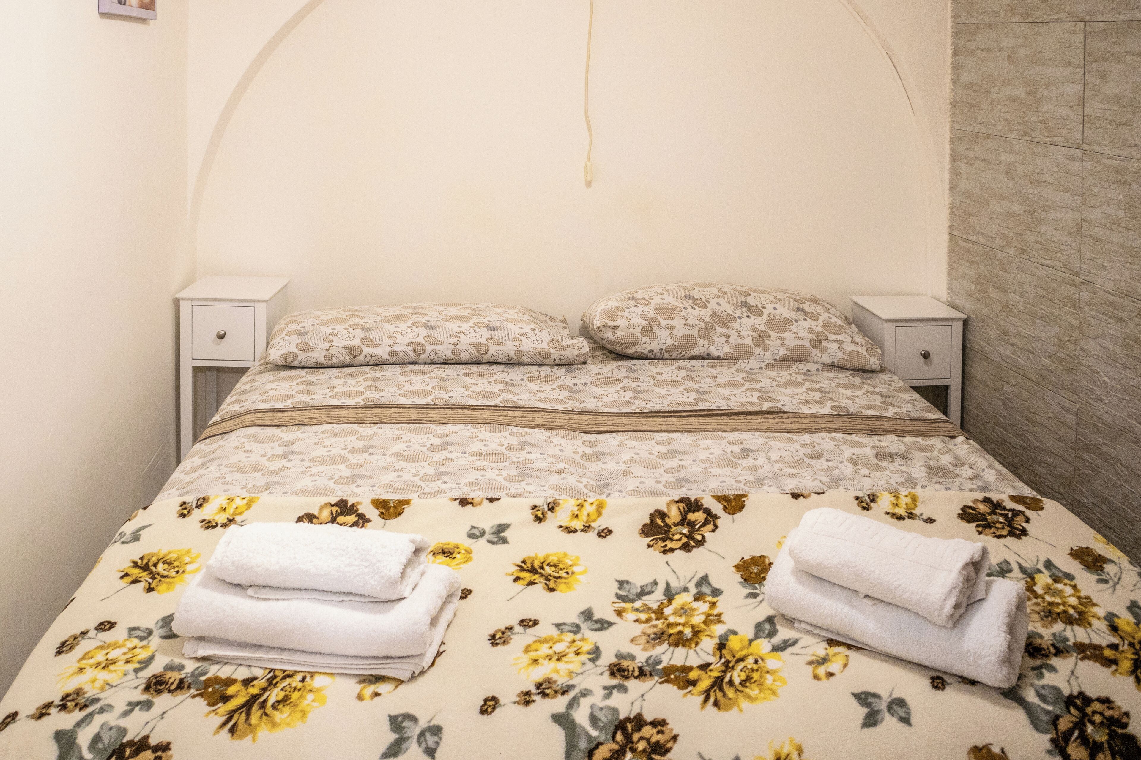 A neatly made bed with floral bedding, two bedside tables, and a wall-mounted power outlet.