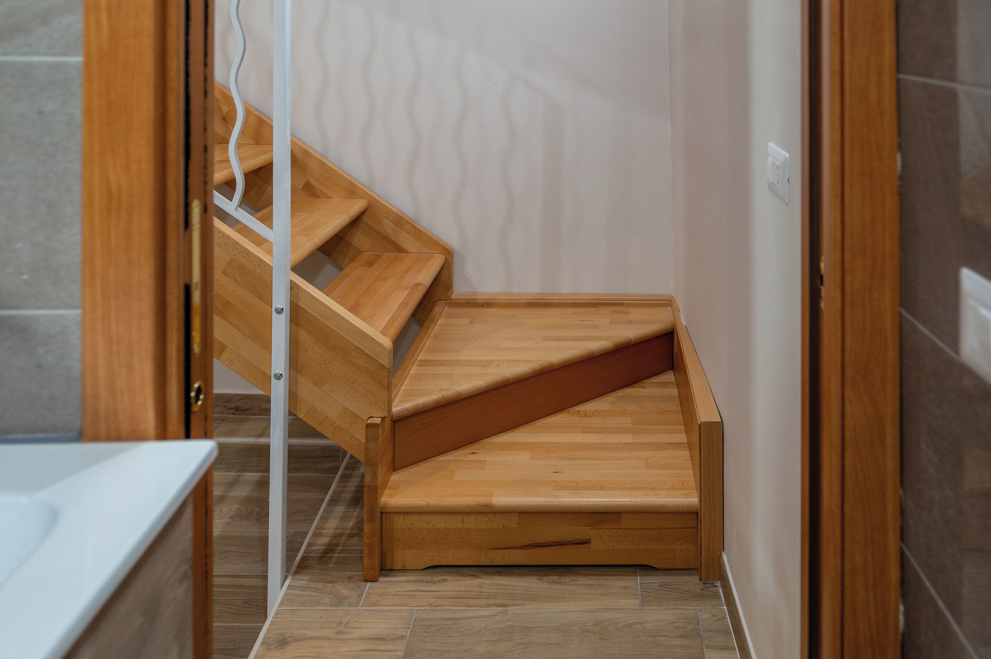 Een houten bank met een gebogen rugleuning in een hoek van een kamer met betegelde vloer en een witte muur.