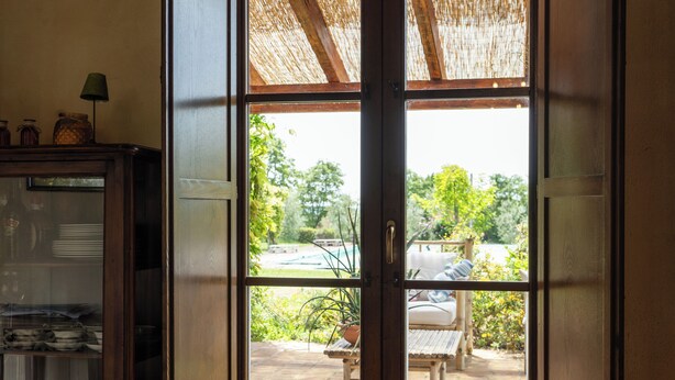 A view through a glass door into a patio area with a wooden table and chairs, a thatched roof, and a garden visible outside. - Casale Farnia - Tenuta di campagna con piscina immersa nel verde (Montecastrilli)