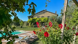 A poolside area with lounge chairs, a house in the background, and a garden with red flowers. - Casale Farnia - Tenuta di campagna con piscina immersa nel verde (Montecastrilli)