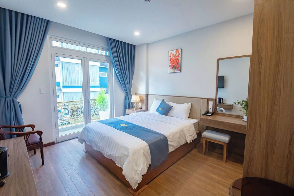 Een hotelkamer met een groot bed, een bureau, een stoel, een kleine tafel, een bankje, een spiegel en uitzicht op het balkon.