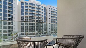 Un balcón con sillas de mimbre y una pequeña mesa, con vistas a una piscina y un edificio moderno de varios pisos.