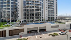 Un edificio moderno de varios pisos con piscina en la azotea, estacionamiento techado y una terraza ajardinada.