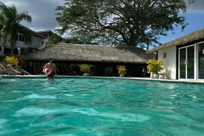 A swimming pool with a person holding a beach ball, surrounded by thatched-roof buildings and potted plants.