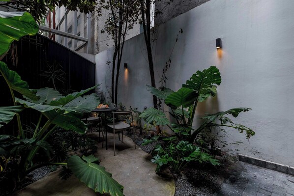 A courtyard with a table and chairs, surrounded by large plants and a wall-mounted light fixture.