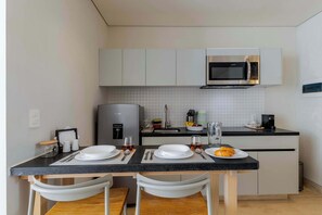 Une cuisine compacte équipée d’un four à micro-ondes, d’une machine à café et d’un évier. On y trouve deux chaises blanches et une table en bois, ainsi que de l’assiette et des couverts.