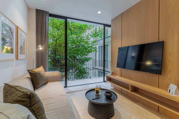 A modern living room with a large flat-screen TV, a sofa with cushions, a coffee table with bottles, and a view through large windows.