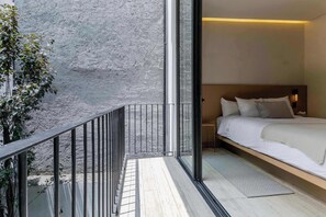 Una habitación de hotel moderna con balcón, cama con ropa de cama blanca, mesita de noche y lámpara de pared.