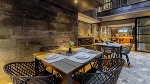 A modern dining area with a stone wall, black wicker chairs, and a long table set for four. - Park Condesa | 2BR, Rooftop Spa & Gym (Ciudad de México)