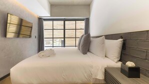 Une chambre moderne avec un grand lit, une tête de lit en bois, une table de chevet avec une boîte de mouchoirs et une fenêtre avec des stores.