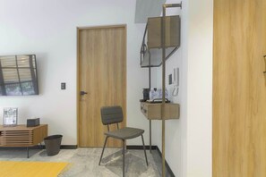 A room with a wooden door, a desk with a chair, a TV mounted on the wall, and a small shelf with items.