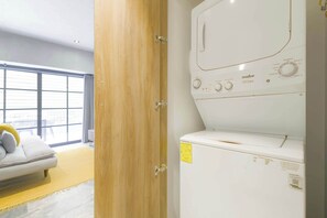 Une buanderie équipée d’un lave-linge et d’un sèche-linge à chargement frontal, d’une porte en bois et donnant sur un espace de vie avec un canapé et une fenêtre.