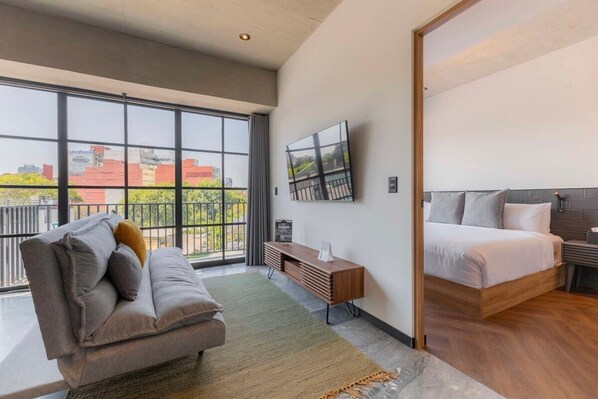 A modern hotel room with a large window, a sofa, a bed, and a TV on the wall.