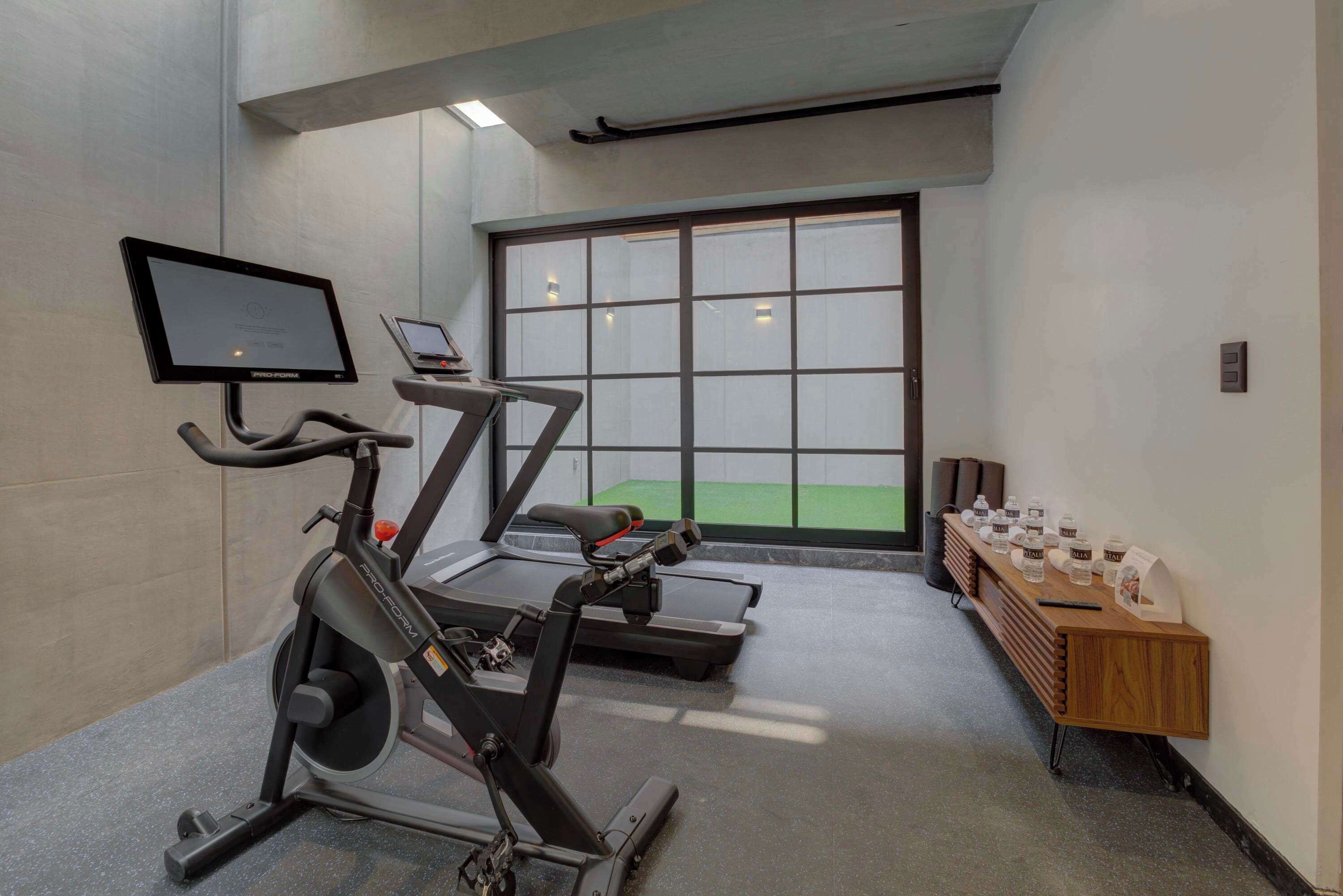Ein Fitnessraum mit Laufbändern, einem Ergometer und einem Rudergerät. Ein großes Fenster bietet Blick auf ein grünes Feld. An der Wand steht ein kleiner Tisch mit Wasserflaschen und einem Schild.