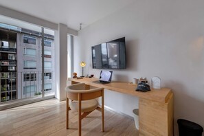 A modern office space with a large desk, a chair, a flat-screen TV, and a view of a building through a large window.