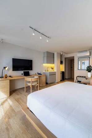 A modern hotel room with a large bed, a desk with a laptop, a TV, and a kitchenette area.