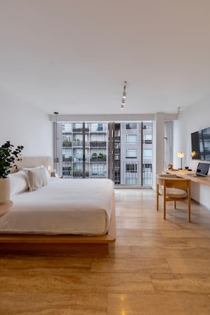 Ein modernes Schlafzimmer mit einem großen Bett, einem Schreibtisch und Blick auf ein Gebäude durch Glastüren.