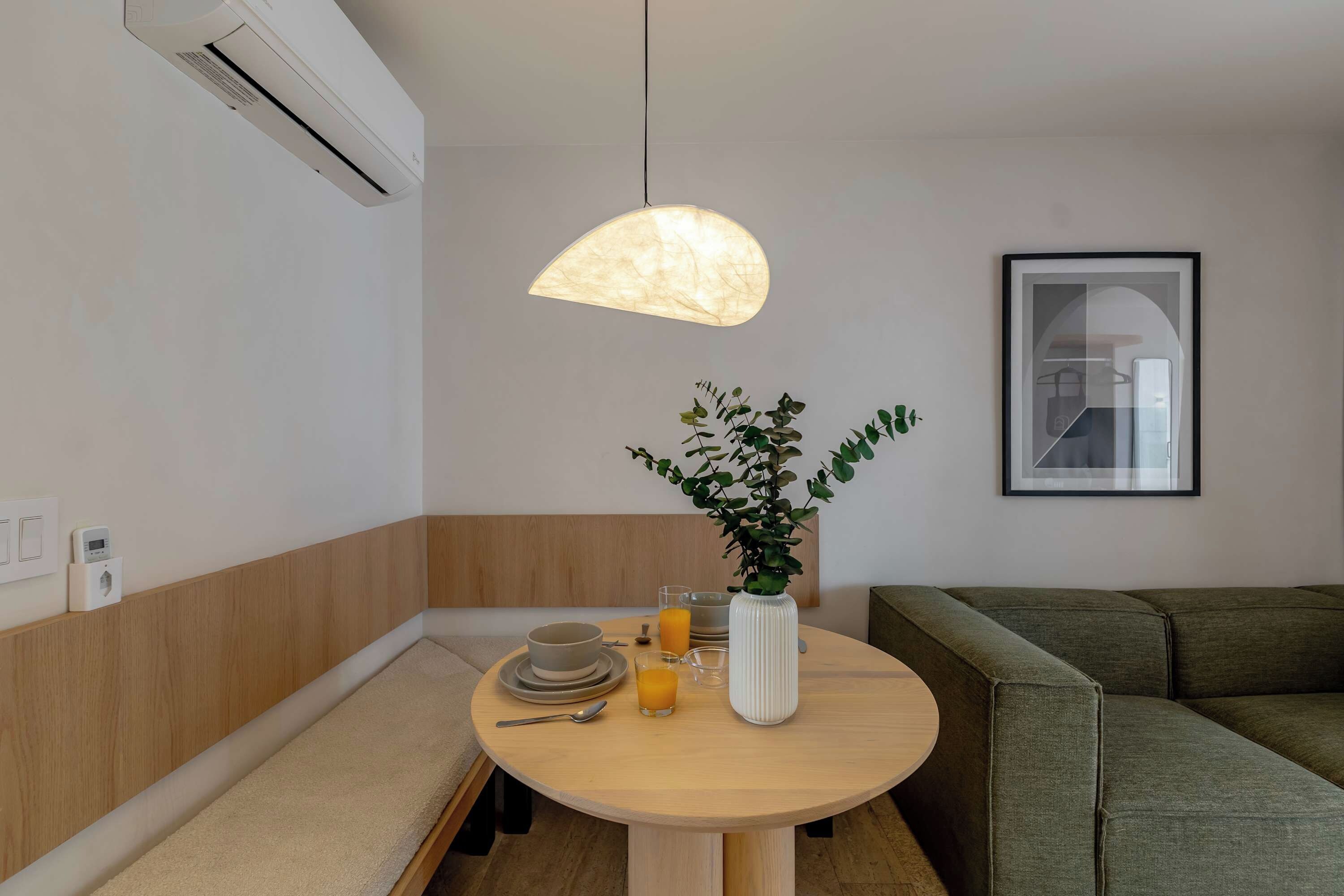 Uma sala de estar moderna com uma mesa de madeira redonda, um vaso branco com folhagem verde e um lustre pendurado.