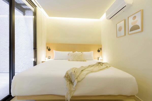 A hotel room with a large bed, an air conditioning unit, a window, and two framed pictures on the wall.