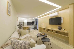 A modern living room with a sofa, two chairs, a small table, a TV mounted on the wall, and a kitchen area in the background.