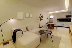 Un salon moderne comprenant un canapé, une petite table ronde, un lampadaire, une plante et un coin cuisine avec un four à micro-ondes.
