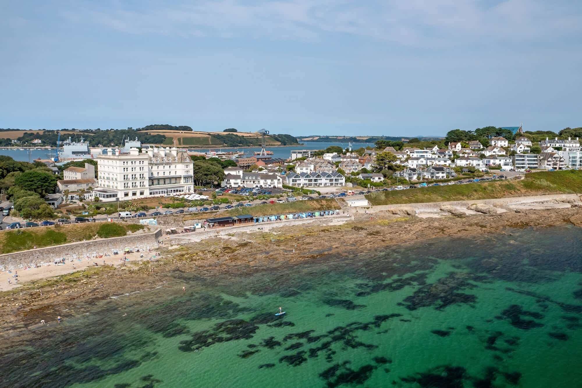 Un hotel costero de varios pisos, una playa en primer plano y vista al mar y colinas distantes.