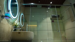 A modern bathroom with a large oval mirror, a white sink, and a glass shower enclosure. - The Infinity Villa (Madikeri)