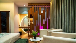 A modern hotel room with a bed, two armchairs, a small round table with a plant, a decorative wall panel, and a view of the outside. - The Infinity Villa (Madikeri)