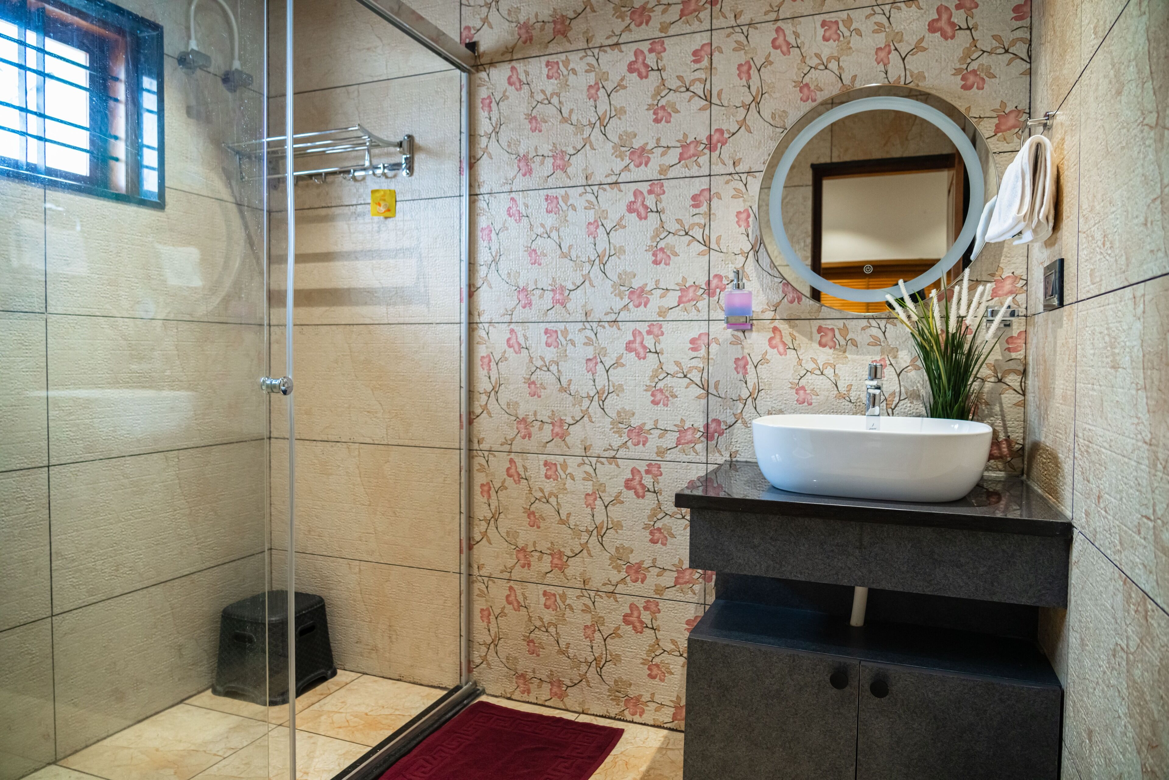 A bathroom with a glass shower enclosure, a white sink with a mirror above it, and floral-patterned tiles.