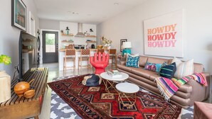 A modern living room with a brown sofa, a red chair, and a patterned rug. There is a dining area in the background with a kitchen visible. - HowdyHouse: Designer Townhouse in Historic FOODIE  Bryan Minutes to Kyle Field (Bryan)