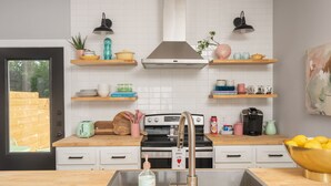 A modern kitchen with white tiles, wooden shelves, a stainless steel sink, and a stove. - HowdyHouse: Designer Townhouse in Historic FOODIE  Bryan Minutes to Kyle Field (Bryan)