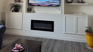 A modern living room with a flat-screen TV mounted on a white wall, a ceiling fan, and a fireplace below the TV. - Located near wineries, tulip festival, outlets, Bauman’s Fram & state parks! (Mount Angel)