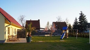 Ein Wohngebiet mit einem Haus, einer Terrasse mit Stühlen, einem kleinen Garten und einem Spielplatz mit einer Rutsche.