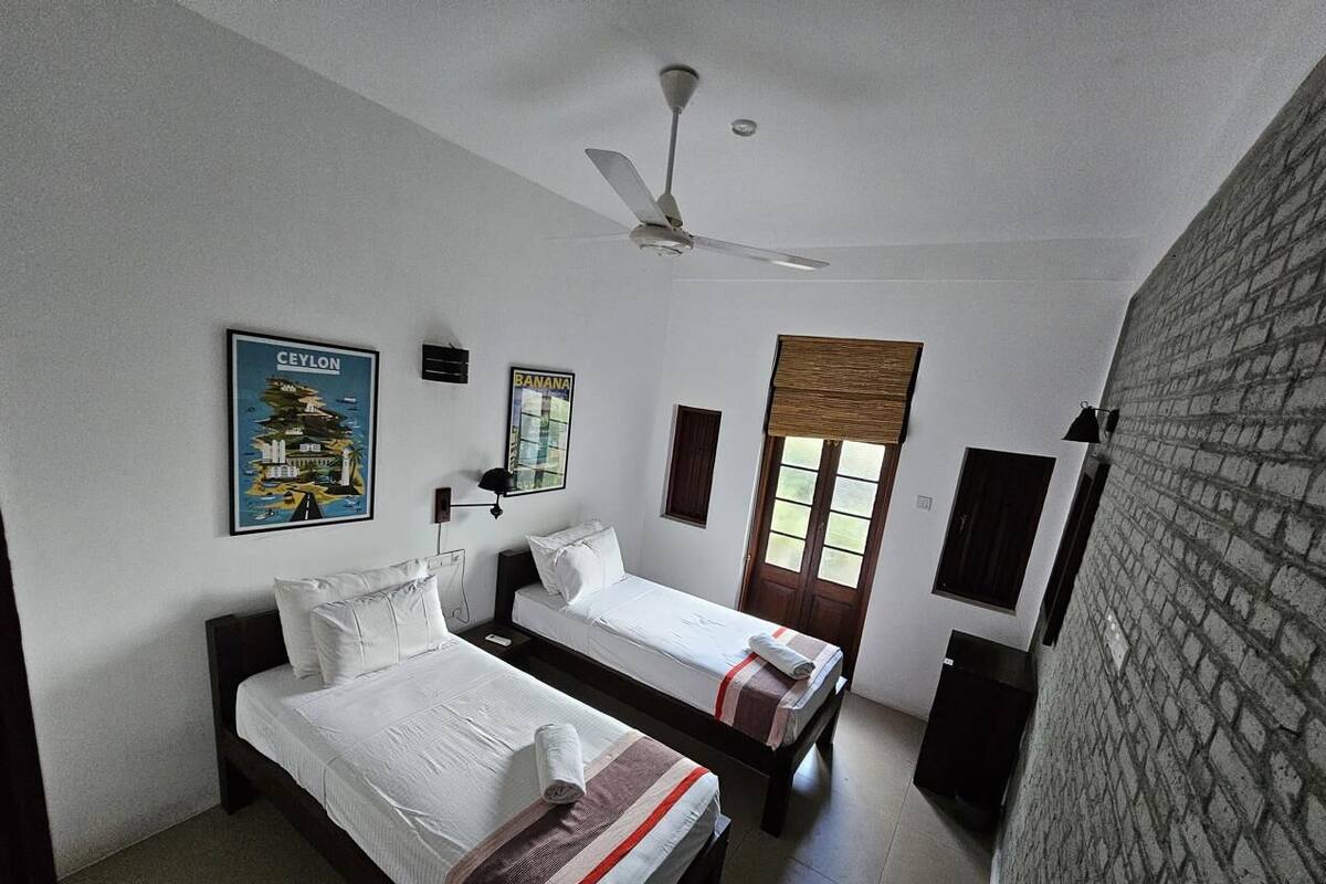 A hotel room with two single beds, a ceiling fan, a window with blinds, and a brick wall.