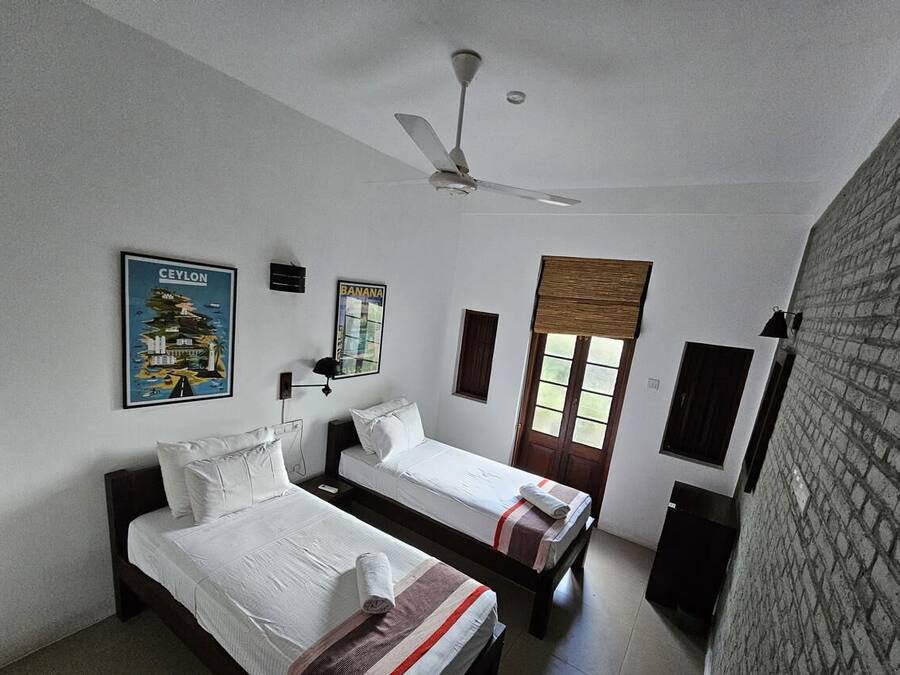 Quarto de hotel com duas camas de solteiro, ventilador de teto, janela com persianas e parede de tijolos.