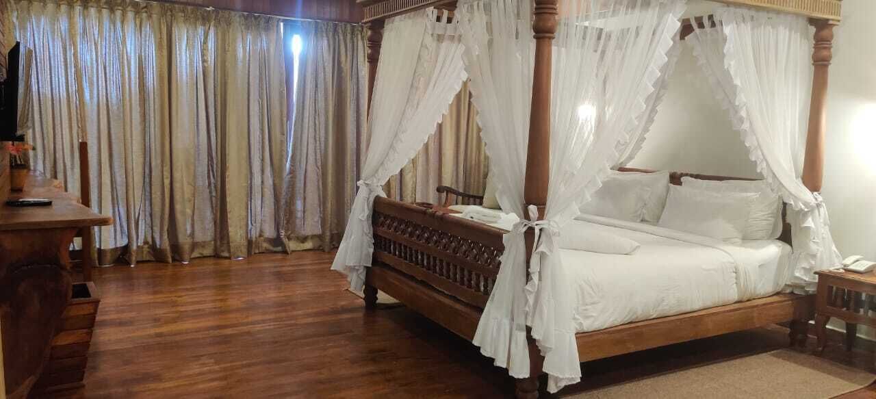 A four-poster bed with white bedding and a wooden frame, a wooden desk with a chair, and a large window with curtains.