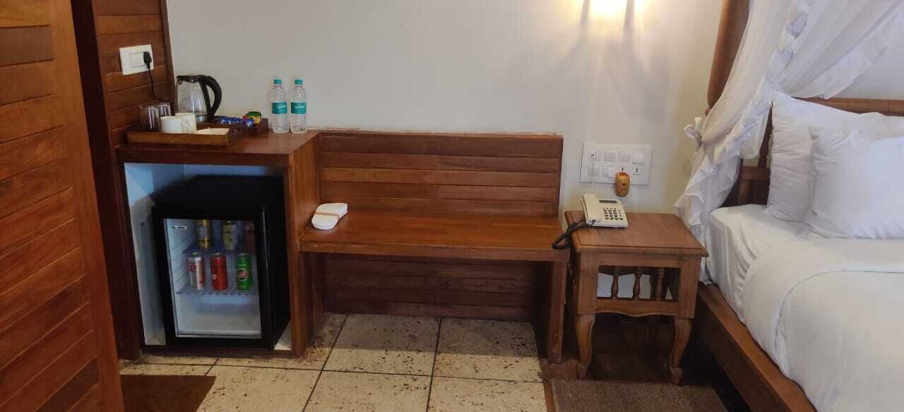 Una habitación de hotel compacta con un banco de madera, un refrigerador pequeño, una mesita de noche con teléfono y una cama con dosel.