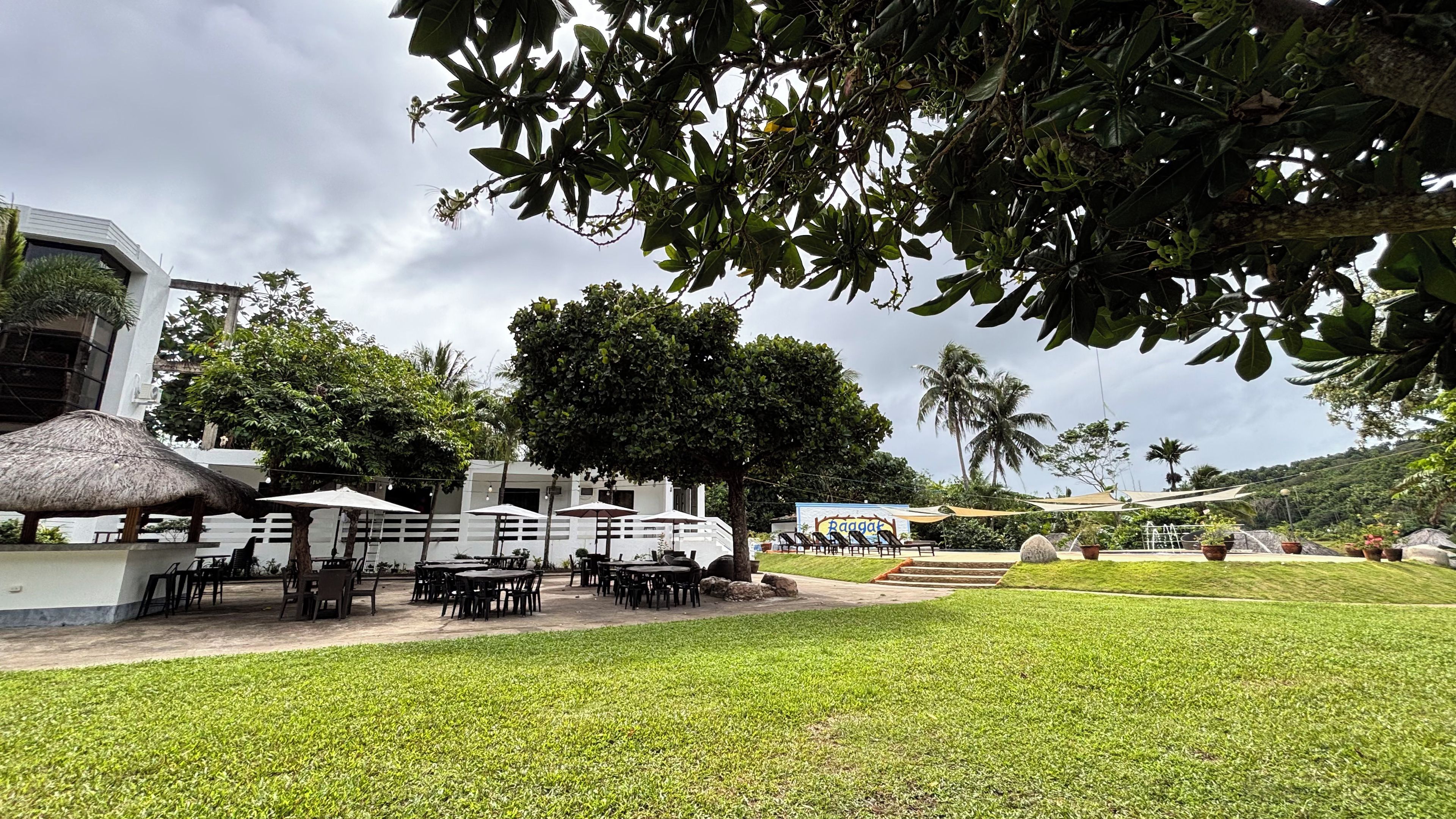 An outdoor seating area with tables and umbrellas, surrounded by greenery and a building with a white facade.