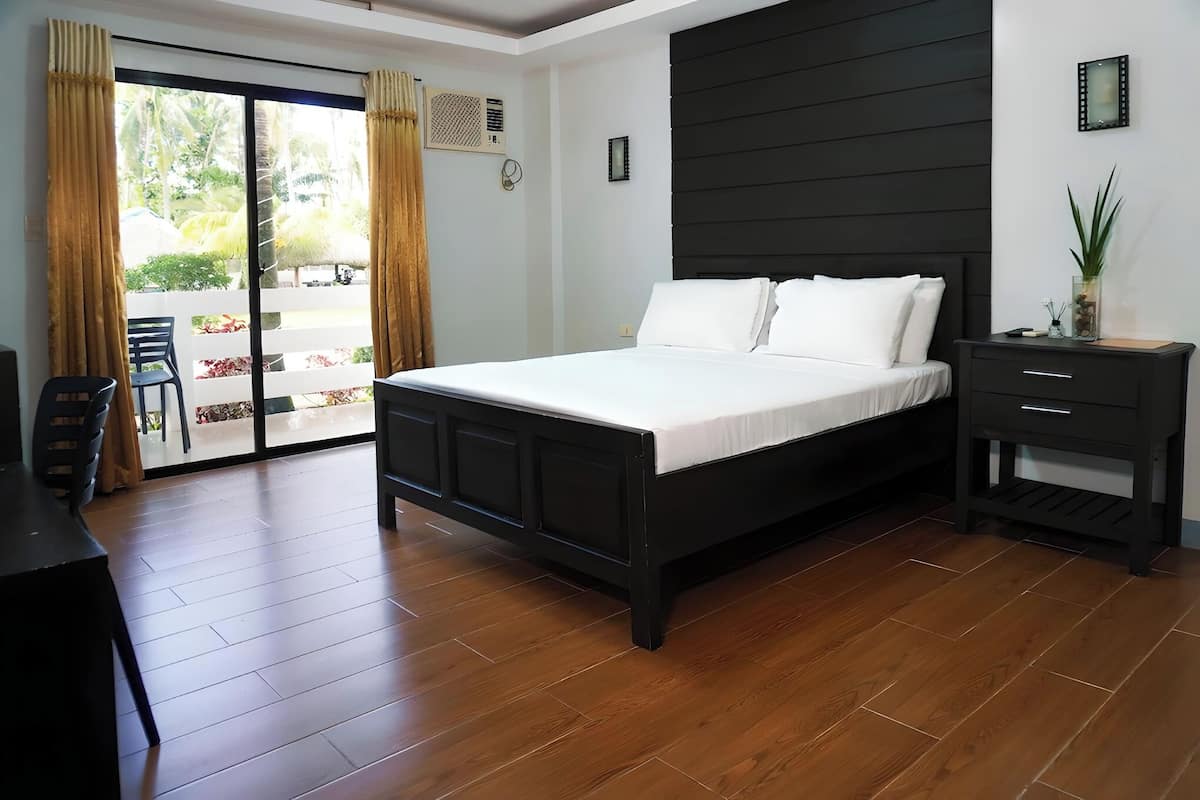 Ein Schlafzimmer mit Bett, Nachttisch, Stuhl und Blick durch eine Schiebetür auf einen Außenbereich mit Pflanzen.