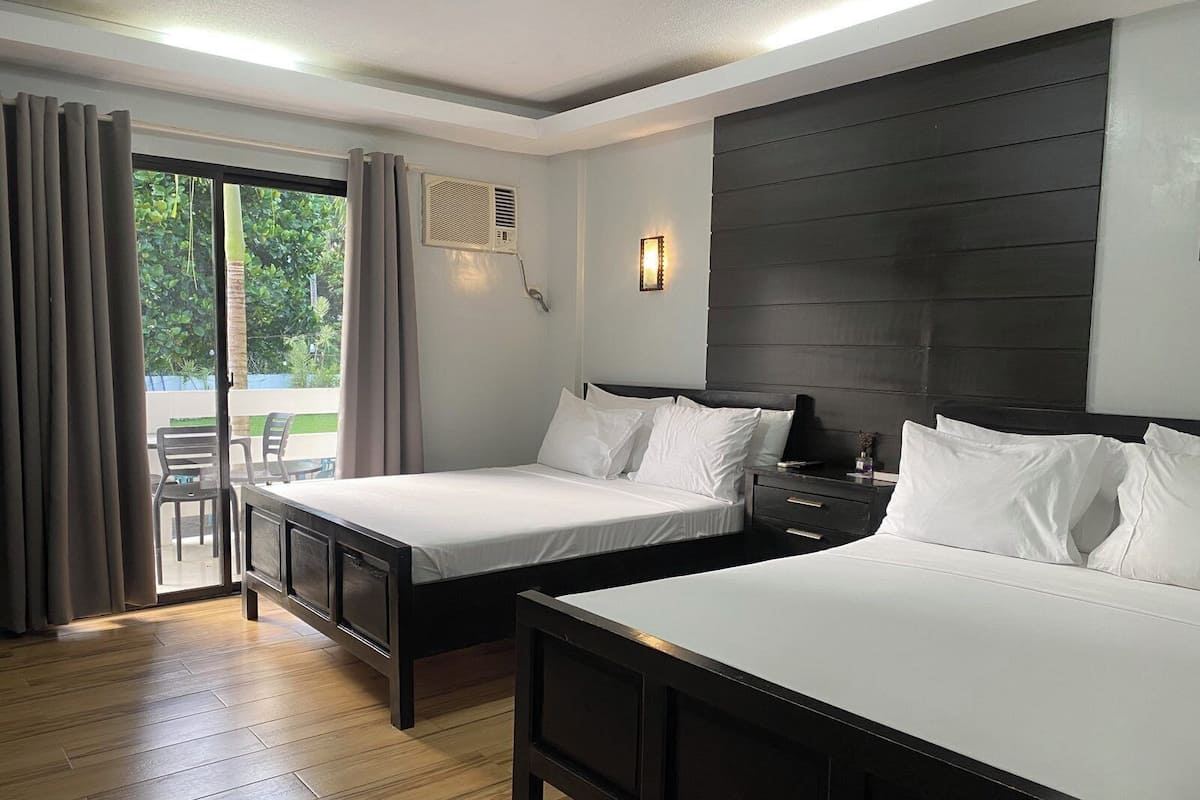 Ein Hotelzimmer mit zwei Betten, einem großen Fenster mit Vorhängen, einem Deckenventilator und Blick ins Freie.