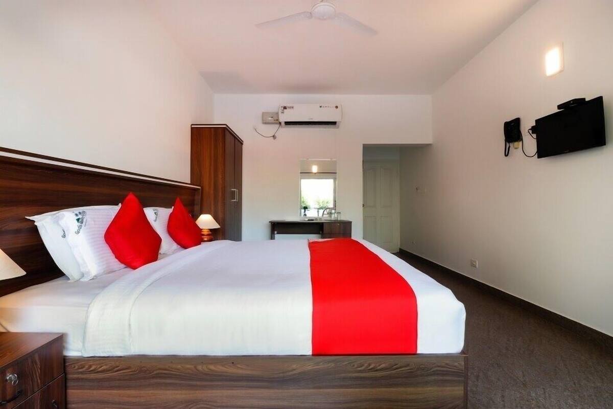 A hotel room with a bed, a desk, a chair, a wardrobe, an air conditioning unit, a television, and a ceiling fan.