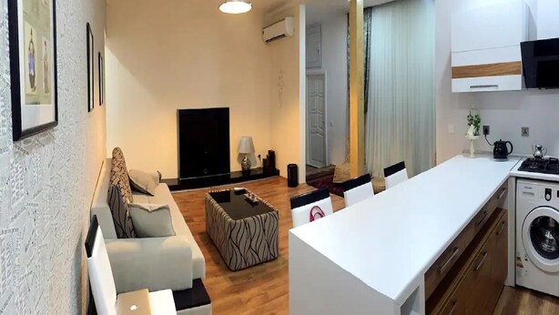 En moderne stue med sofa, sofabord og tv. Der er et køkkenområde med komfur og vaskemaskine.